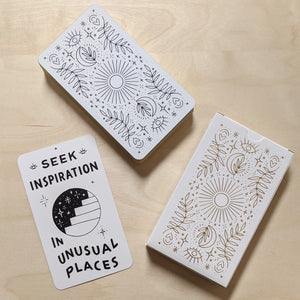 (clockwise from bottom left corner) white oracle card with black drawing of a starry staircase portal reads "seek inspiration in unusual places", stacked card deck features gilded edges & magical symbol motifs in black, deck box featuring magical symbol motifs in gold by Worthwhile Paper available at Coyote Supply Co, a zero waste witch store in Midtown Reno, Nevada that is BIPOC owned