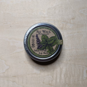 Round metal tin with round kraft paper label featuring a green boarder & a sprig of mint containing vegan lip balm by Sea Witch Botanicals available at Coyote Supply Co, a zero waste witch store in Midtown Reno, Nevada that is BIPOC owned