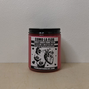 Red soy wax spell candle in a clear glass jar with a black lid.  Black & white label reads "Como La Flor, lovely lover come to me, sweetness, amor, attraction, love" & features art of 3 roses & an anatomically correct heart.  Made by Firme Arte & sold by zero waste witch store Coyote Supply Co in Reno, Nevada.