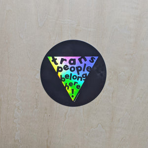 Round black sticker features a rainbow holographic triangle in the center that reads "trans people belong here!" in black text.  By Ash & Chess, sold by zero waste witch store Coyote Supply Co in Reno, Nevada