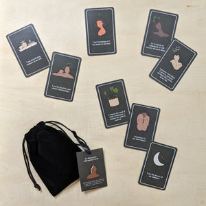 (clockwise from bottom left)  Black velvet drawstring pouch with a paper hang tag featuring a portrait of the artist, Ashley, a black woman with dark shoulder length hair.  8 black paper cards are scattered above the deck, randomly, each card features a drawing & a phrase.  The drawings are of a variety of humans, all different skin tones & gender presentations, all have sprouts growing from the tops of their heads.  Sold by zero waste witch store Coyote Supply Co in Reno, Nevada.