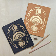 Load image into Gallery viewer, Black and terracotta six month open dated planners by the Rainbow Vision. Both covers feature a gold embossed rainbow with a moon and sun bordered by snakes. Below the notebooks are a wooden pencil sharpener and two wooden pencils available at Coyote Supply Co, a zero waste witch store in Midtown Reno, Nevada that is BIPOC owned