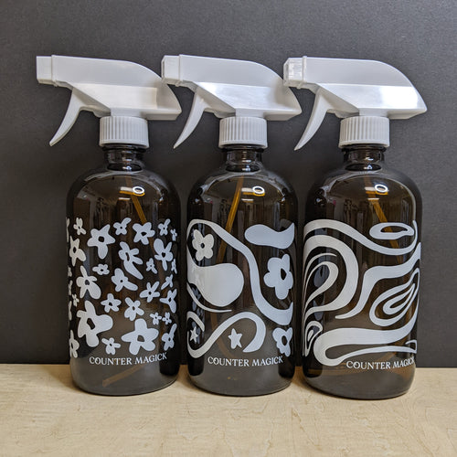 Three 16oz dark amber glass bottles that say Counter Magick in white text, and white plastic spray tops. Each bottle sports a different design from left to right they feature white flowers, white flowers and stars with swirls, and white swirls.  Sold at Coyote Supply Co a BIPOC and LGBTQIA+ owned zero waste witch store in Reno Nevada