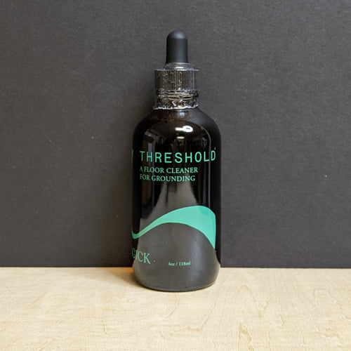 A 4oz  dark amber glass bottle with a dropper top that reads: Threshold A Floor Cleaner For Grounding in green text.  Sold at Coyote Supply Co a BIPOC and LGBTQIA+ owned zero waste witch store in Reno Nevada