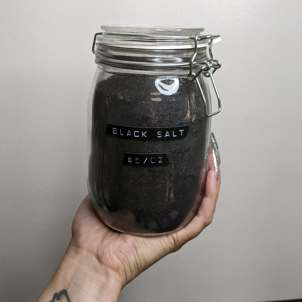 A glass jar full of black salt being held up by a hand against a grey background. The jar has a black label with white text that reads: Black Salt $5/oz. Sold at Coyote Supply Co a BIPOC and LGBTQIA+ owned zero waste witch store in Reno Nevada