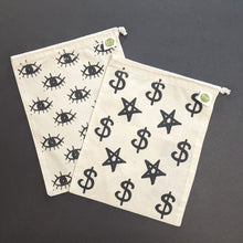 Load image into Gallery viewer, Two beigie cotton drawstring bags. The left one has a black ink design of many eyes screenprinted on. The right bag features five pointed stars and dollar signs in black screenprinted on. Sold at Coyote Supply Co a BIPOC and LGBTQIA+ owned zero waste witch store in Reno Nevada