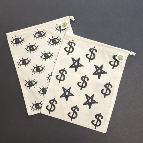 Two beigie cotton drawstring bags. The left one has a black ink design of many eyes screenprinted on. The right bag features five pointed stars and dollar signs in black screenprinted on. Sold at Coyote Supply Co a BIPOC and LGBTQIA+ owned zero waste witch store in Reno Nevada