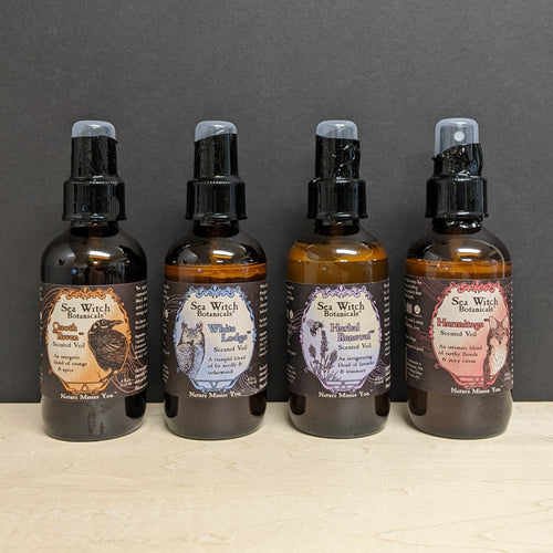 Four amber glass bottles on a black background. The bottles each has a black label with a colored frame in the center that says Sea Witch Botanicals Scented Veil. From left to right the labels are 
orange with an illustration of a raven Quoth the Raven, blue with an illustration of a great horned owl White Lodge, purple with an illustration of a bee on lavender Herbal Renewal, red with an illustration of a fox Hermitage.
Sold at Coyote Supply Co a BIPOC and LGBTQIA+ owned zero waste witch store in Reno NV