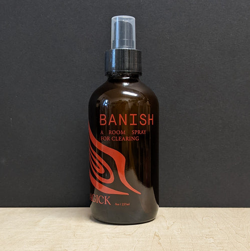 An 8oz dark amber glass bottle with a spray top that reads: Banish A Room Spray For Clearing in red text. Sold at Coyote Supply Co a BIPOC and LGBTQIA+ owned zero waste witch store in Reno Nevada
