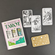 A light purple box that reads Color Your Own Tarot in green text, the box also features images of the High Priestess card in black and white and various states of color. Next to the box are uncolored black and white versions of The Tower, The Fool, and the Knight of Swords. Below the cards is the back of a card, it is light brown with white stars, moons, and suns. All on a black background.  Sold at Coyote Supply Co a BIPOC and LGBTQIA+ owned zero waste witch store in Reno Nevada