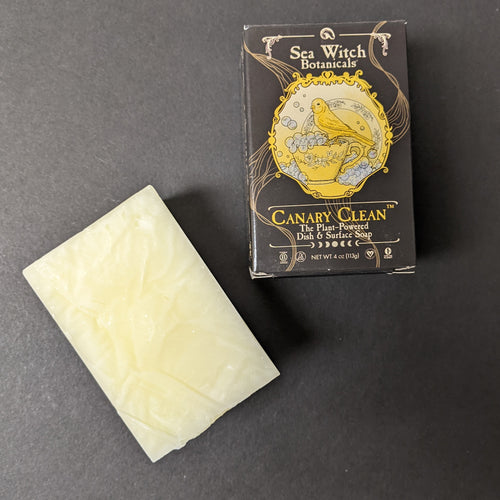 A light yellow bar of canary clean next to it's black box on a black background. The box has an illustration of a canary on bubbly dishes, the label reads: Sea Witch Botanicals 
CANARY CLEAN
The Plant-Powered
Dish & Surface Soap